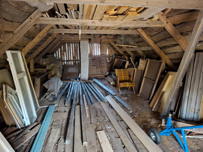 Gammalt uthus med träbjälkar, fönster uppställda, plankor och verktygslåda. Planerat för ombyggnad till verkstad och ungdomsbostad.