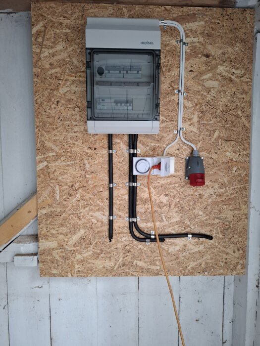 Elcentral monterad på spånskiva i en vägg, med ledningar och en röd elkontakt bredvid.