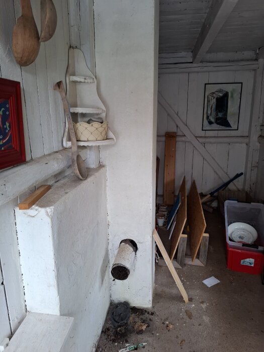 Gammalt uthus med vitmålade väggar, hyllor och verktyg. Röret in i väggen antyder tidigare användning. Inredning och renoveringsmaterial syns.