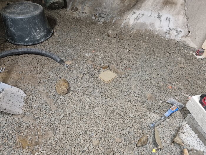 Byggarbetsplats med grusgolv, svarta hinkar, verktyg och slangar. Stenvägg i bakgrunden.
