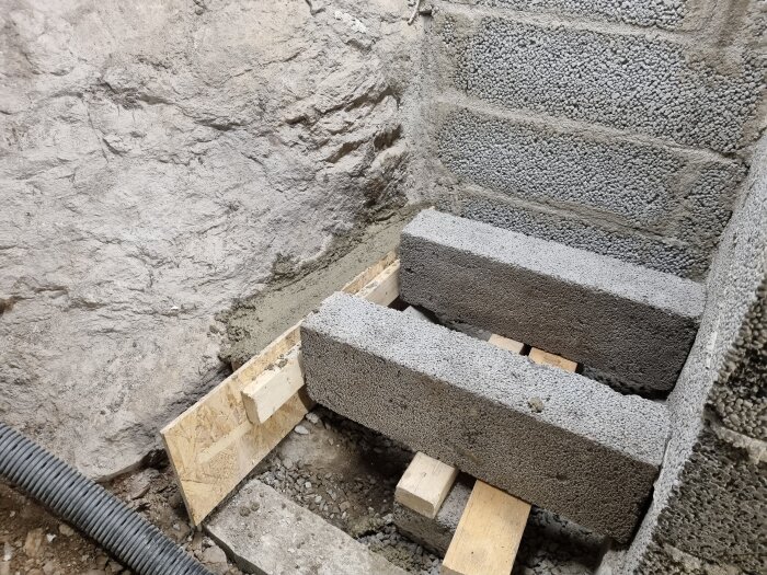 Betongblock staplade i hörn bredvid en grov stenvägg med träbjälkar och formgjutning; synlig mark och rör vid sidan.