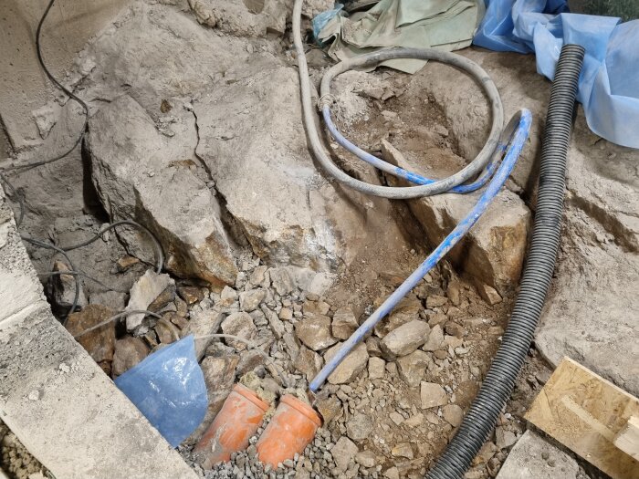 Byggarbetsplats med exponerad sten och jord, flera kablar och rör, samt orange plastdetaljer insprängda i marken.