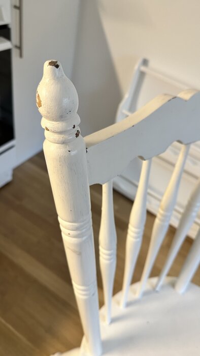 Sliten vit stol med avskalad färg på ryggstödets knopparm och pinnar, står på ett trägolv.
