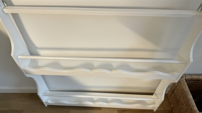 Vit hylla med två våningar och rundade detaljer, monterad mot en vägg ovanför en träkorg.