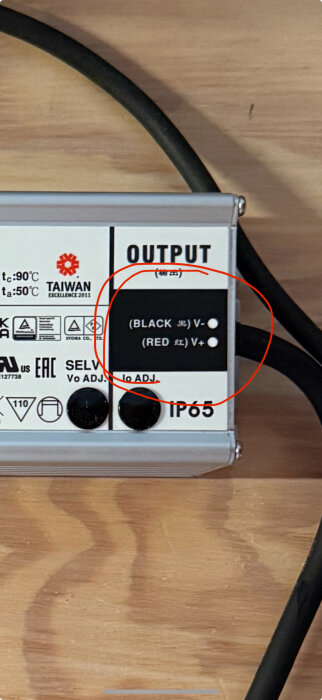 Närbild av en transformator med text som visar sekundärsidan: svart V-, röd V+, med omgivande märkning och etiketter.