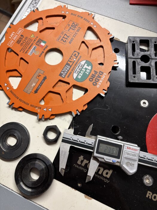 En orange dadoklinga, distansringar och en skjutmått som visar 29,97 mm ligger på ett arbetsbord för träbearbetning.