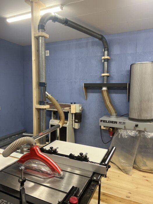 Verkstad med nyinstallerad spånsugsrörledning, sågmaskin och dammutsug i ett blått rum med träbjälkar och arbetsbänkar.