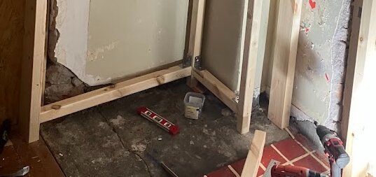 Byggprojekt med reglar monterade direkt på ett stengolv i ett rum, omgivna av verktyg som vattenpass och borrmaskin.