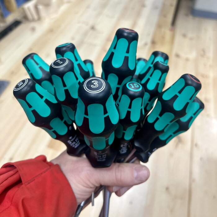Hand som håller en bukett av skruvmejslar med gröna och svarta handtag på ett trägolv.