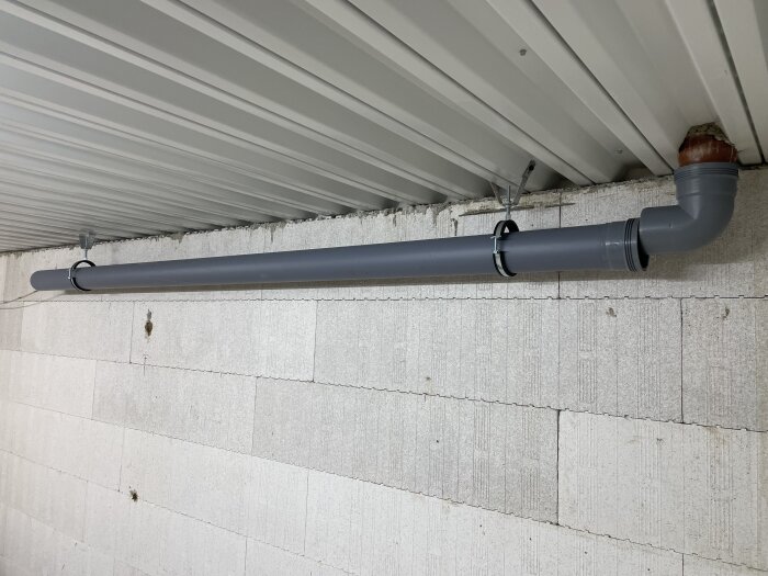 Grått avloppsrör monterat i taket med lutning, fastsatt med metallklämmor, förberett för senare anslutning till avloppssystem.