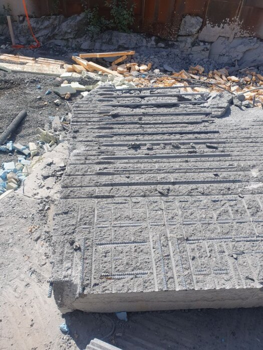 Betongplatta med synliga armeringsjärn, delad i två delar på en byggarbetsplats, omgiven av bråte och träspillror.