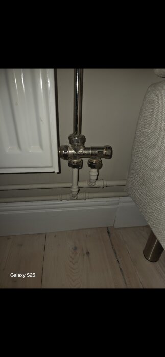 Närbild av en radiatorventil med rörkopplingar vid ett golv, delvis synlig radiator och parkettgolv i bakgrunden.