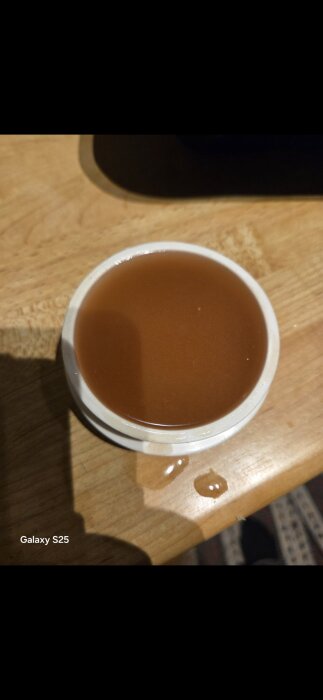Brun vätska i en vit kopp på en träyta, med några droppar spillda bredvid.