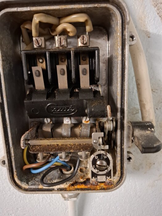 Närbild av en elektrisk kopplingsbox med olika färgade kablar och komponenter, delvis rostig och smutsig.