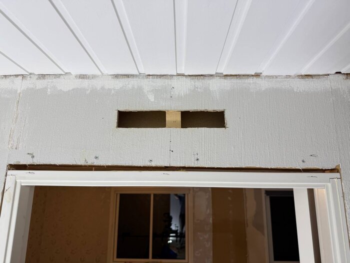 Öppning för överluftsdon ovan dörr, synlig vertikal träregel kvar i innervägg, del av bärande konstruktion, tak av vita panelbrädor ovanför.