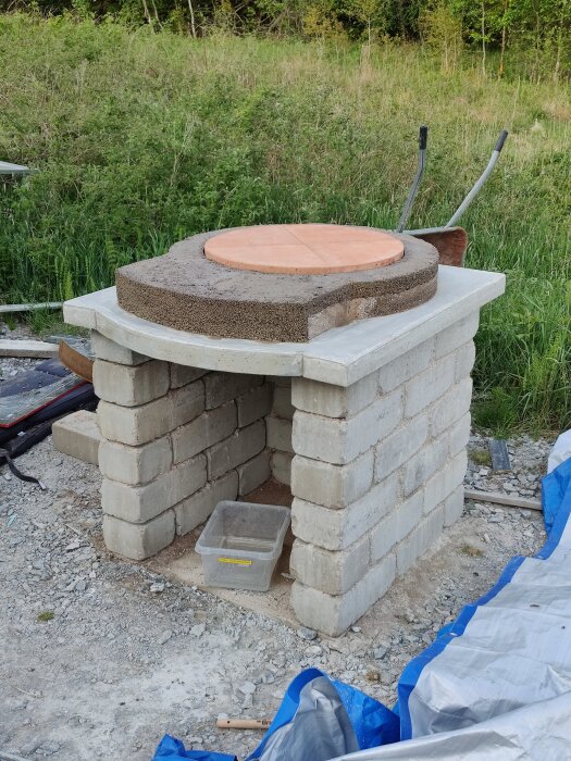 Utomhusbyggd stenugn under konstruktion med ett trätäcklock ovanpå cementbas och lämningar av byggmaterial runt.