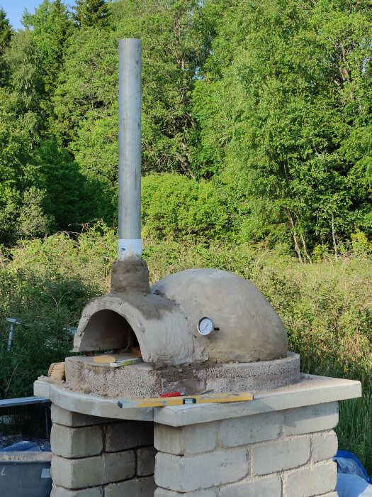 Vedeldad ugn av lera och stenull med skorsten, byggd i trädgårdsmiljö, med tegelbas och termometer på ovansidan.