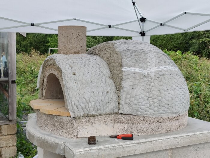 Ugn under konstruktion med valv av betong, isolerad med nät och puts, placerad utomhus på stenbas med verktyg bredvid.