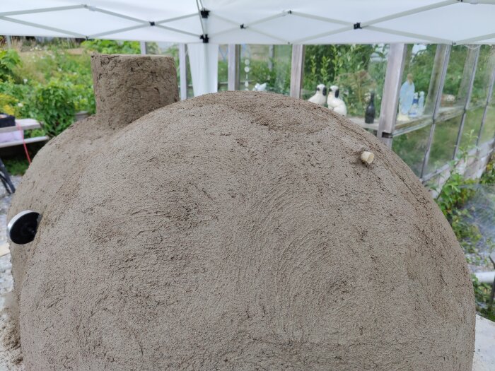 Utomhusbild av en hembyggd ugn med runda former och rustikt utseende, omgiven av växter och ett tält.