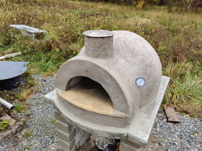 Hemmagjord lerugn med termometer och skorsten, byggd på sandgrund. Ugnens väggar av homebrew, stenull och puts. Står på grusig mark.
