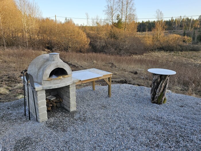Utomhus vedeldad ugn byggd av betong och tegel med vedförvaringsutrymme, placerad på grusad yta vid träbord och stubbe.