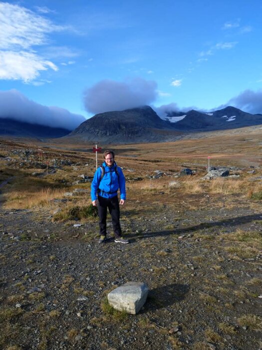 En person i blå jacka står på en stenig stig i ett fjällandskap med berg och moln i bakgrunden.