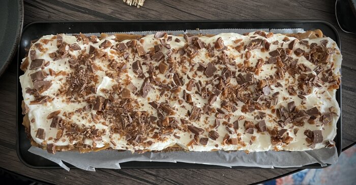 Kladdkaka toppad med kolasås, skivad banan, grädde och hackad daim på en mörk plåt, med en botten av krossade mariekex.