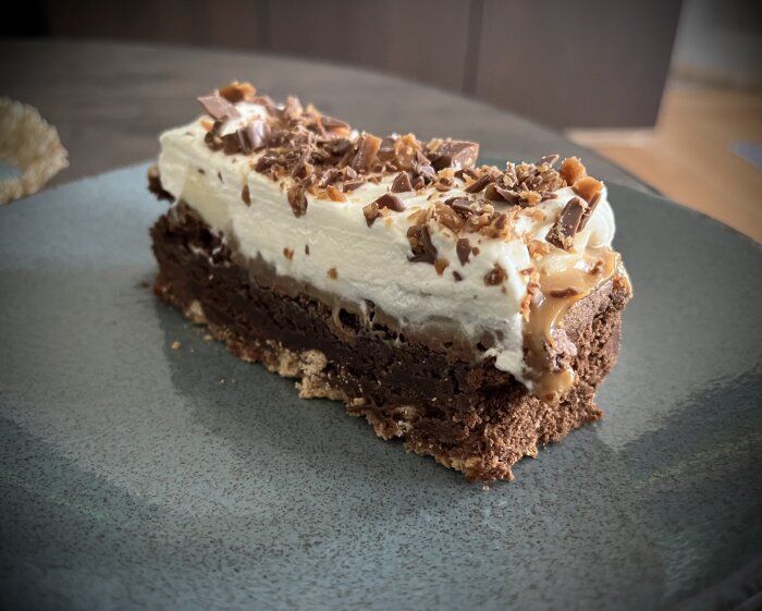 Banoffee och kladdkaka-kombination med mariekex-botten, chokladkaka, kolasås, skivad banan, grädde och hackad Daim på svart tallrik.