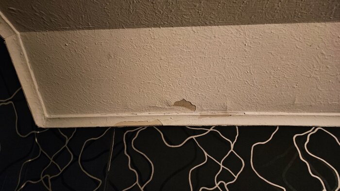 Flagande vit färg på innertak nära murad yta, med ojämn textur och svarta tapeter med linjemönster i förgrunden.