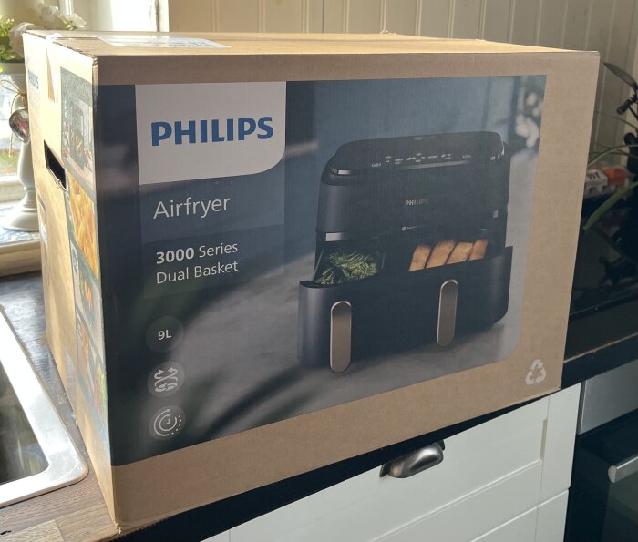 Kartong med Philips Airfryer 3000 Series Dual Basket, 9 liter, på en bänkskiva framför ett fönster.