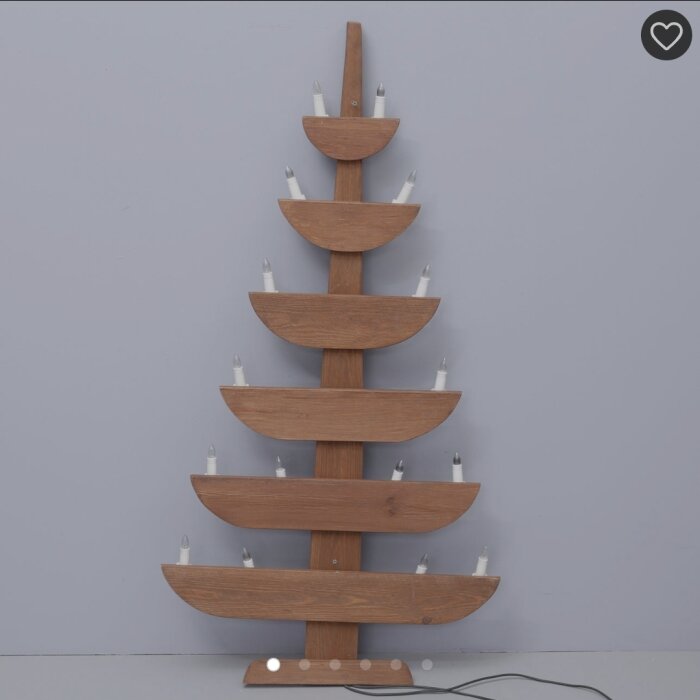 En träjulgran med ljushållare och glödlampor placerad mot en grå vägg.