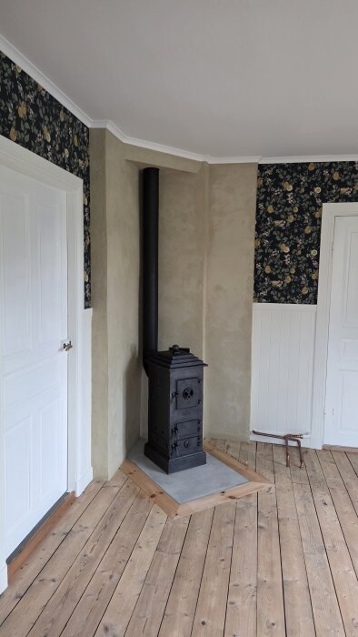 Kamin i hörn av rum med träpanel och mönstrad tapet, golv av träplankor och närliggande dörr.