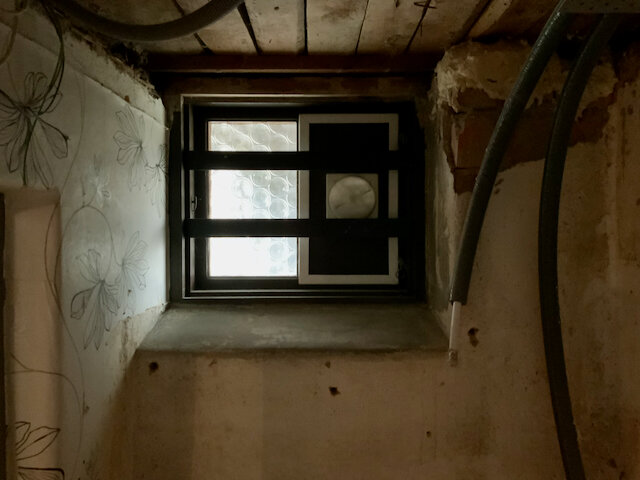 Ventilationsfönster med mörk ram och blomtapet runt, kanalrör syns till höger i en odekorerad vägg till ett badrumsprojekt.