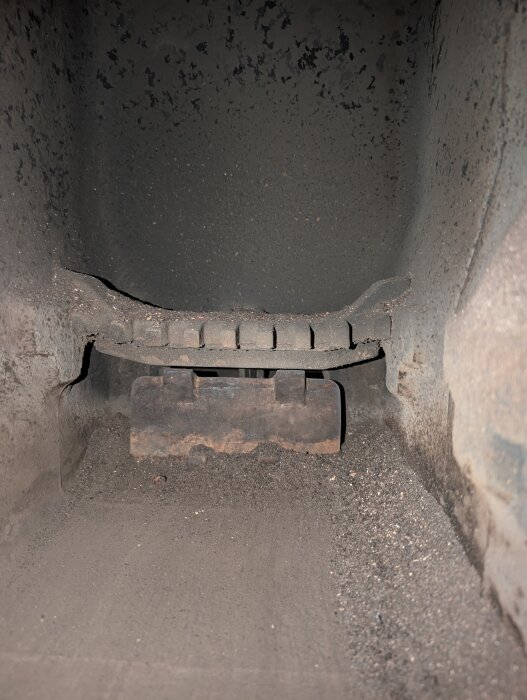 Inuti en kamin eller ugn med rostigt galler syns en metallplåt och aska.