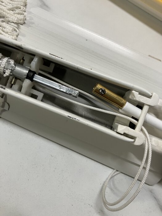 Vridstångsmekanism med flexvajer och guldfärgad hylsa i en persienn, monterad med låsskruv, på vit bakgrund.