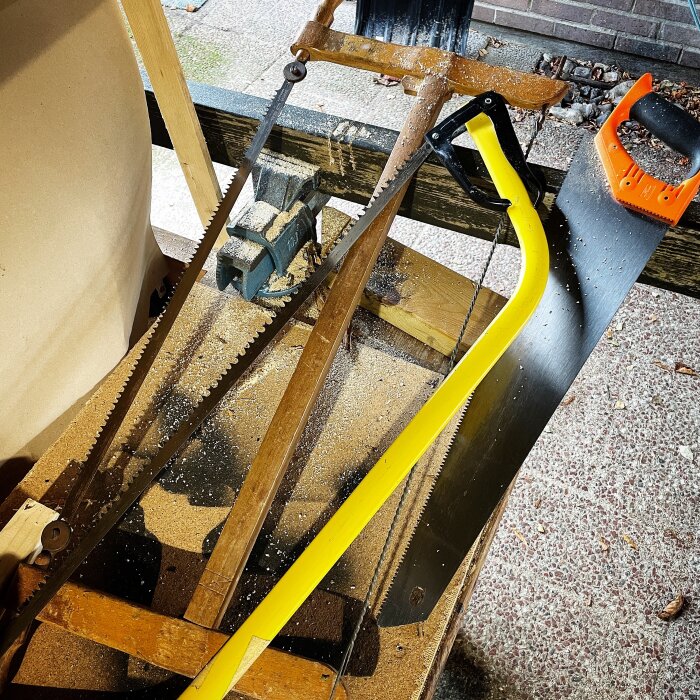 Tre olika typer av sågar ligger på ett arbetsbord med sågspån: en i trä, en med gul båge och en modernt orange handtag.
