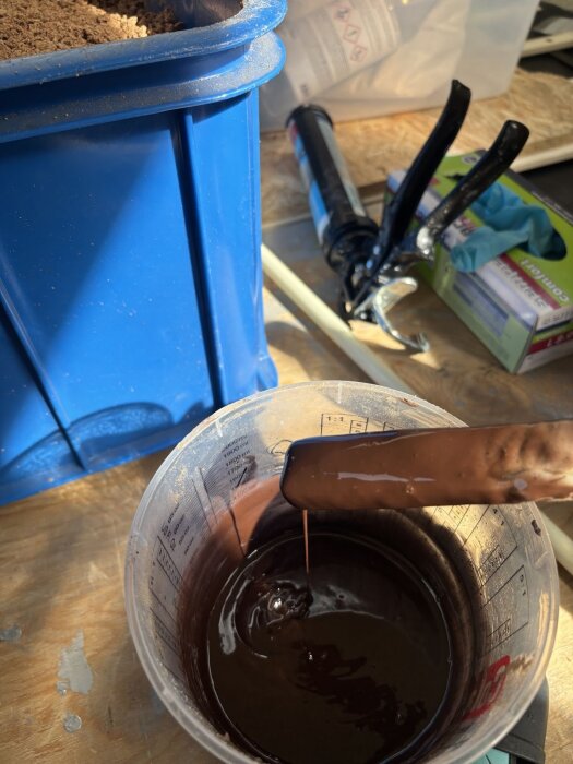 Brun blandning av kork och silikon i en mätbägare, bredvid en blå behållare och en fogpistol.