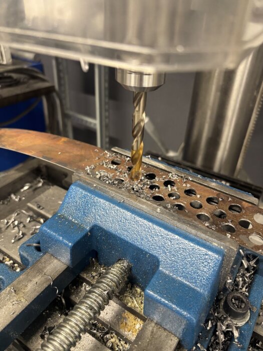 Borrmaskin som används för att borra hål i en plåtbit, placerad i ett skruvstycke med metallspån runtom.
