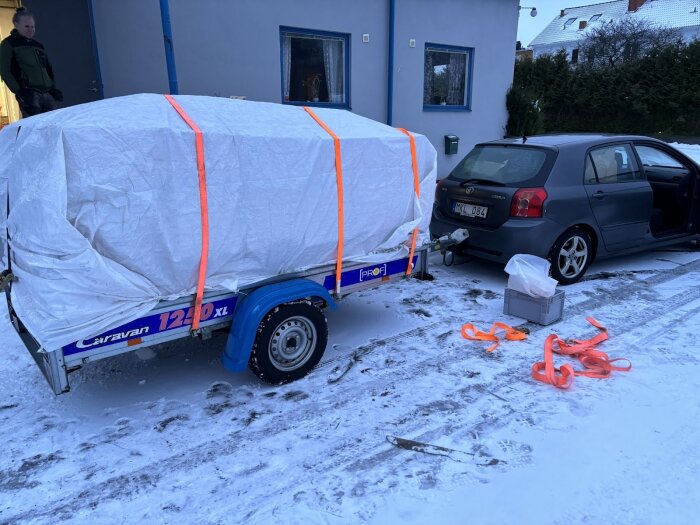 Bil med släpvagn lastad med täckt material, står parkerad vid ett hus. Snö på marken och lösa spännband på sidan.