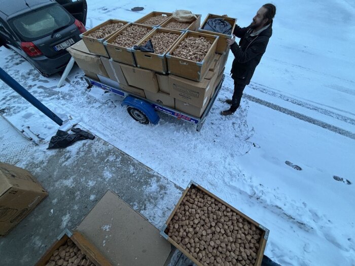 En man lastar lådor fulla med korkbitar på ett släp under vintertid. Det ligger snö på marken och en bil står parkerad i bakgrunden.