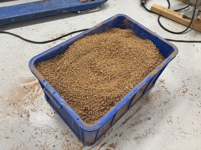 Blå plastlåda fylld med grovkornigt mald kork placerad på ett grått betonggolv, med korkspån utspridda runtomkring.