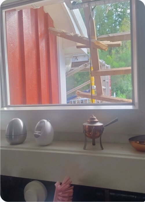 Utsikt genom fönster visar byggställning utanför hus med röd fasad. Insidan har äggformade objekt och en liten kopparkastrull.