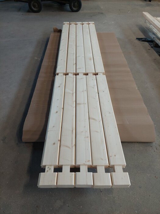 Plankor för bastubygge i trä, lagda på brun papp på golvet, med urfrästa snitt i ändarna för att visa olika byggeriondknutar.