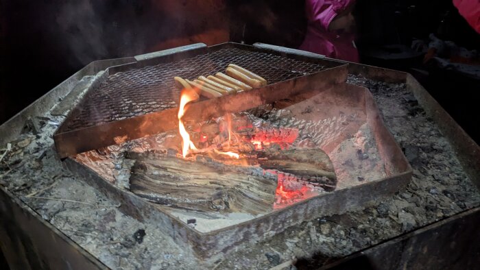 Korvar som grillas över en öppen eld på ett metallgaller i en eldstad.