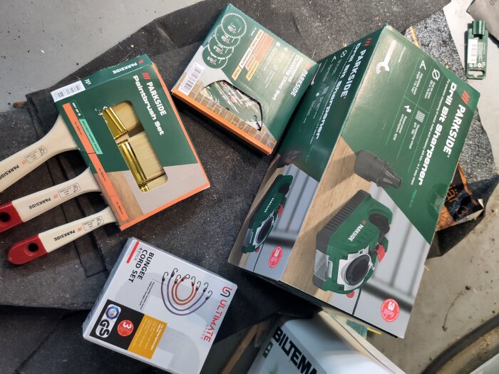 Förpackningar med en PARKSIDE borrslip, penselset och ULTIMATE spännbandsset ligger på ett bord eller golv.