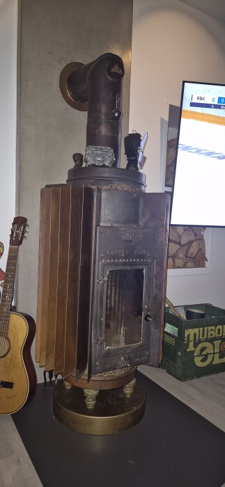 Gammal kamin märkt "Copper King" med glaslucka, omgiven av gitarr och trälåda i ett vardagsrum. På väggen hänger ett TV-spel.