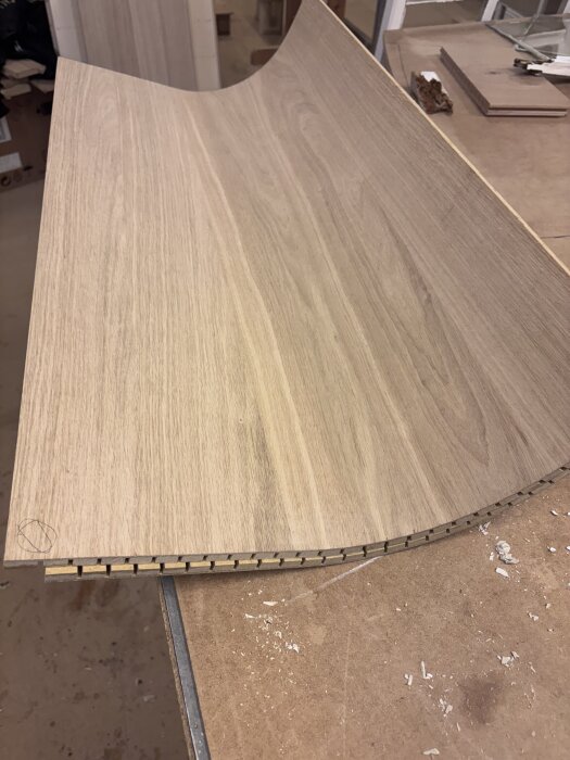 Svängd trälucka med fasad kantlist under bearbetning på en arbetsbänk, synliga träspån och verktygsdelar i bakgrunden.