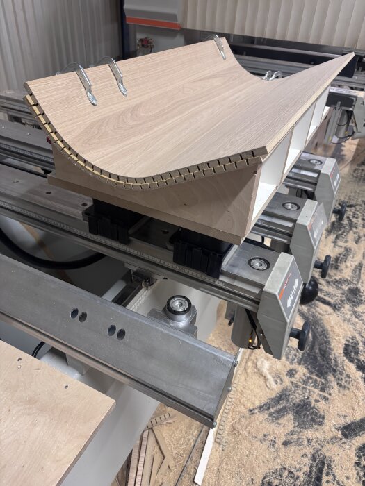 Svängd träbit klämd i metallstöd på arbetsbord, omgiven av sågspån. Används för att skapa en fasad kantlist.