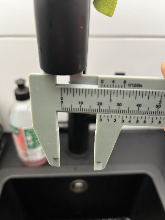 Närbild på en mätning med skjutmått av utloppsröret på en köksblandare; bakgrunden visar en diskho och en tvålpump.