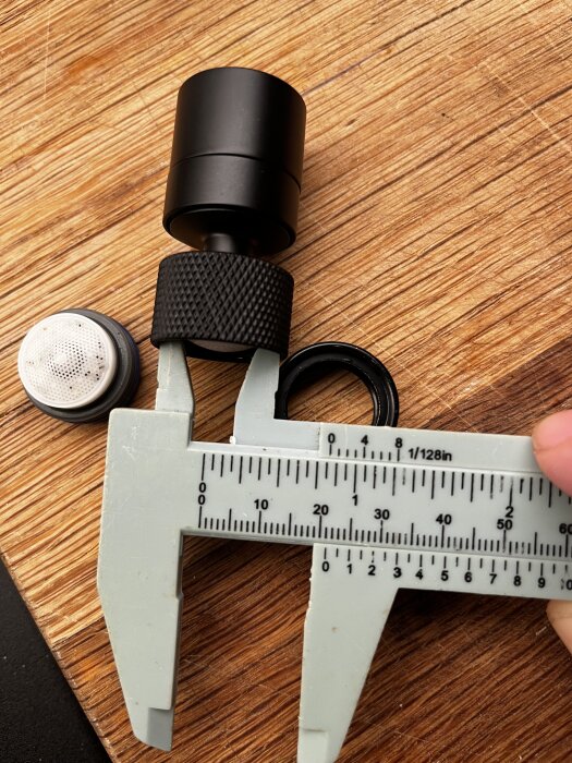 Svart strålsamlare, borttagen från en blandare, placeras bredvid en skjutmått på en träyta. En vit filterdel syns också.